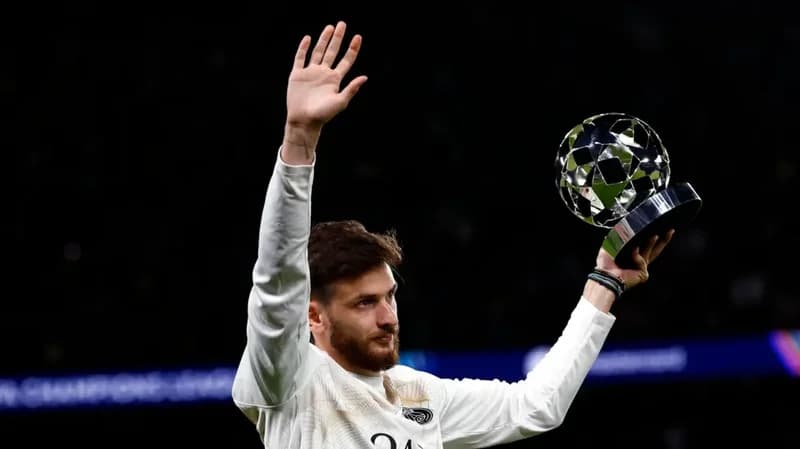 Georgian footballer Khvicha Kvaratskhelia is captured waving to a crowd while holding a silver star-patterned trophy, likely a Player of the Match award. He is wearing a white long-sleeved jersey against a dark stadium backdrop with blurred lights.