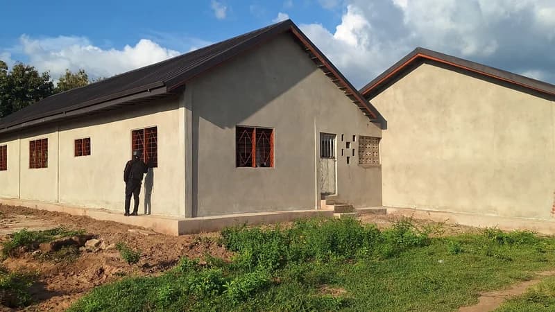 A newly constructed school building with grey plastered walls and a dark corrugated roof under a clear sky. A person in dark attire stands near a window with brown security bars, and the surrounding area shows fresh soil and green vegetation.