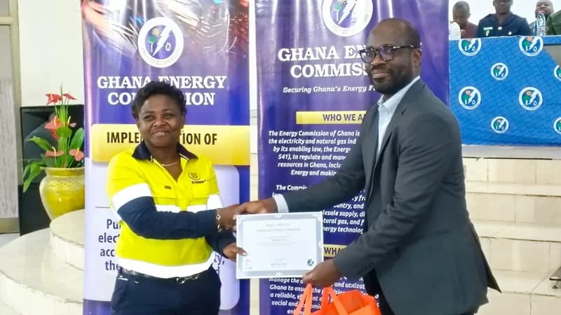 A man in a formal suit presents a certificate to a woman wearing a high-visibility yellow and navy blue work uniform during an official Ghana Energy Commission event. They are shaking hands and smiling in front of commission banners and a flower arrangement.