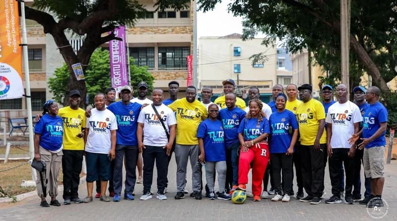 A large group of staff and academics from Accra Technical University pose together during the TUTAG Week celebrations. Participants are wearing branded t-shirts in yellow, blue, and white, with one woman in the foreground resting her foot on a soccer ball, suggesting a sports or wellness activity.