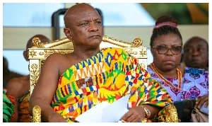Togbe Afede XIV, the Agbogbomefia of the Asogli State, is seated on an ornate golden throne while wearing a vibrant traditional Kente cloth. He is holding a white document and appears to be attending a formal ceremony, with other attendees visible in the background.