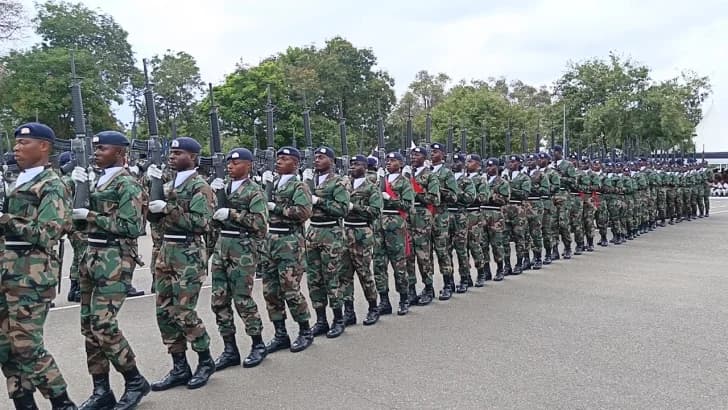 A large contingent of Ghana Army soldiers march in a disciplined formation during a military parade. Dressed in woodland camouflage uniforms and blue berets, the soldiers carry rifles and wear white ceremonial gloves against a backdrop of trees and an overcast sky.