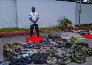 A man stands behind an array of seized items displayed on the ground outdoors. The display includes various firearms, ammunition belts, camouflage uniforms, and tactical equipment, likely evidence from a police operation.