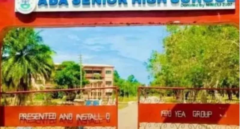 The photograph depicts the main entrance gate of Ada Senior High School in Ghana, featuring a prominent red metal structure. Behind the gate, campus buildings and tropical trees are visible under a bright sky.