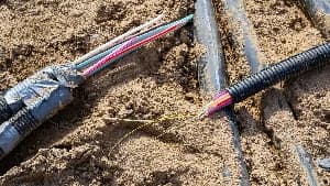 A photograph showing damaged underground telecommunications infrastructure. Severed fiber optic cables with exposed multi-colored wires are visible inside black corrugated conduits within a sandy trench.