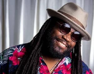 A portrait of reggae musician Gramps Morgan, smiling and wearing a beige fedora and dark round sunglasses. He is dressed in a dark blue floral shirt with large pink hibiscus patterns, positioned against a background of white curtains.