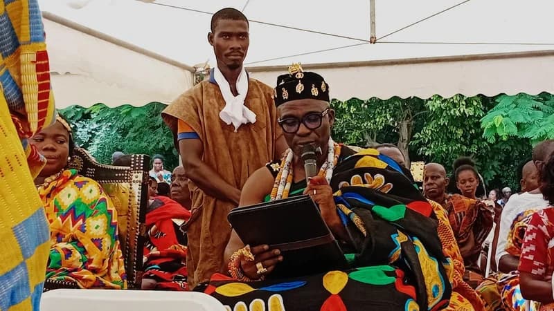 A Ghanaian chief dressed in vibrant traditional Kente cloth and intricate gold ornaments reads from a folder while speaking into a microphone. He is seated under a white canopy during a formal outdoor ceremony, accompanied by attendants and community members.