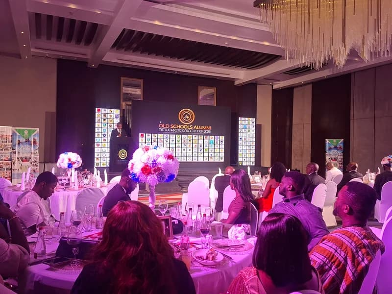 A formal gala dinner for the Old Schools Alumni Networking event held in a decorated ballroom. Guests are seated at round tables with white linens facing a stage where a speaker stands at a podium in front of a large digital screen displaying numerous school crests.