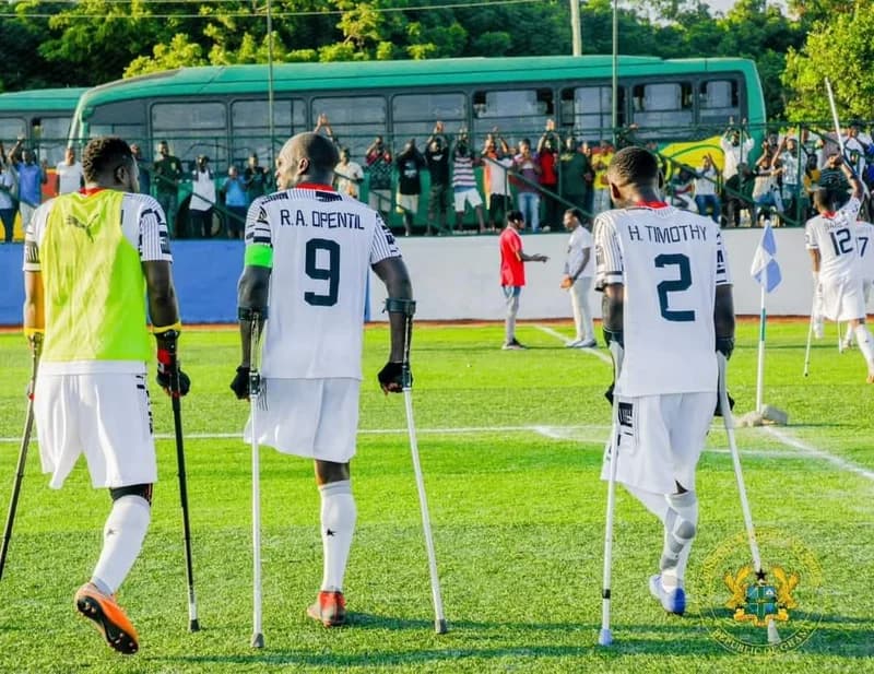 The image shows members of Ghana's national amputee football team, the Black Challenge, walking on a green pitch. Three players are prominently featured from behind wearing white jerseys with black text and using crutches for support, with spectators and a green bus visible in the background.