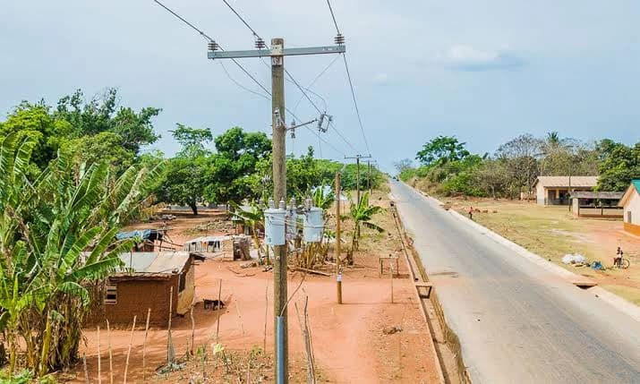 A photograph of a paved road in a Ghanaian community featuring electricity infrastructure including a wooden utility pole with a transformer and overhead power lines. The scene is surrounded by lush green vegetation and simple residential structures, representing rural infrastructure development.