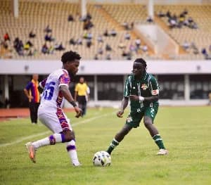 A Karela United player in a green kit challenges a Hearts of Oak player wearing a white jersey with purple and red accents during a Ghana Premier League match. The action takes place on a grass pitch with a backdrop of a stadium filled with yellow seating and spectators.