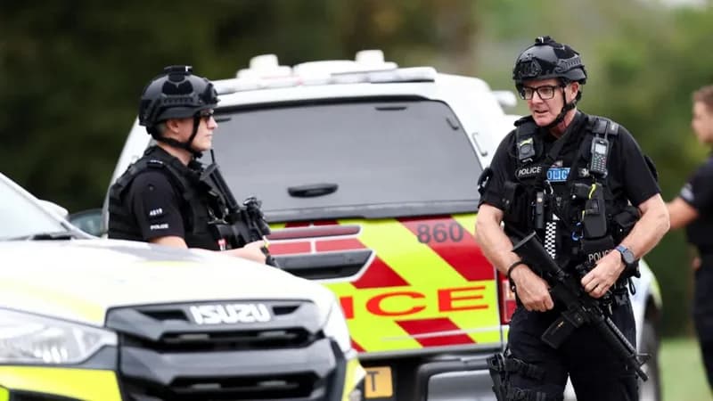 Armed police officers in tactical gear are pictured standing near marked police vehicles. One officer in the foreground carries a rifle while another stands behind an Isuzu vehicle with high-visibility yellow and red markings, indicating a high-security operation.