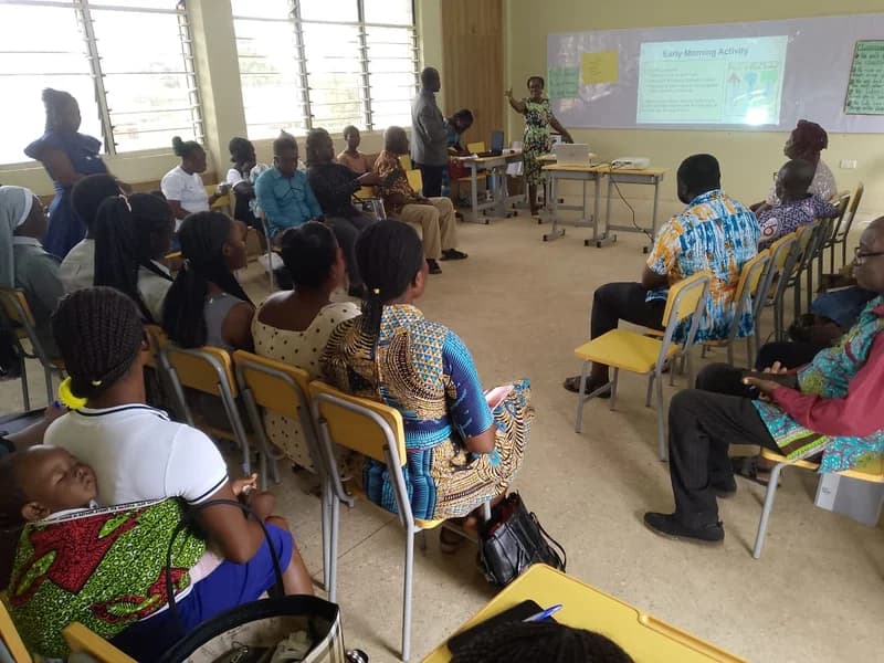 A group of educators attends an in-service training session for Early Childhood Development (ECD) in a classroom. A female facilitator at the front points to a projection screen displaying 'Early Morning Activity' while participants listen attentively.