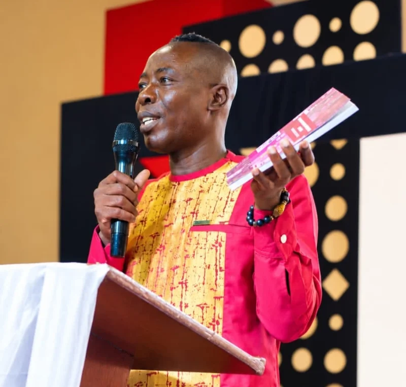 A photograph of Prof Kwofie speaking at a podium, holding a microphone and a book. He is dressed in a vibrant red and yellow traditional Ghanaian shirt and appears to be giving a presentation at a conference or workshop.