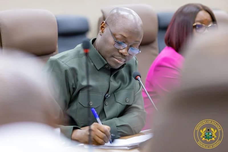 Dr. Cassiel Ato Forson is captured in a focused moment, wearing glasses and a dark green shirt while writing during an official meeting. The setting includes microphones and a Ministry of Finance seal, suggesting a formal government or economic discussion.