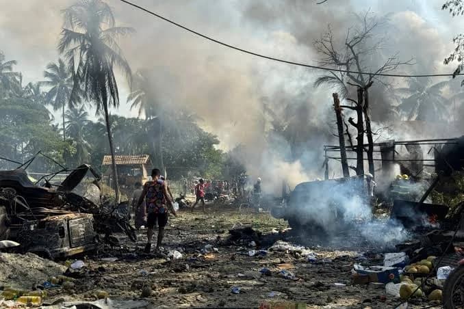 A scene of devastation following an airstrike shows thick smoke rising from charred wreckage and debris. People are seen navigating through the rubble and destruction in a tropical setting with palm trees in the background.