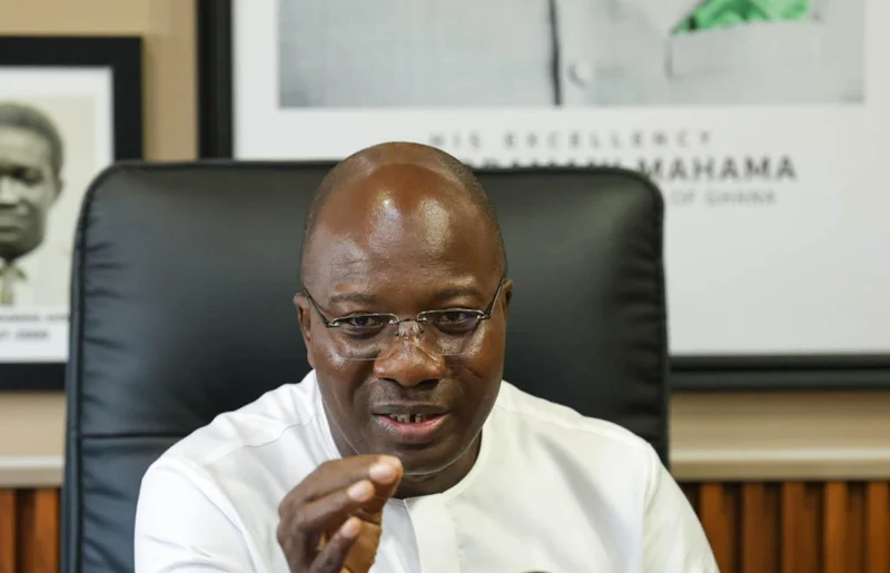 A close-up photograph of a Ghanaian official wearing a white shirt and glasses, gesturing with his hand while speaking. In the background, an official portrait of President John Dramani Mahama is visible on the wall, indicating the setting is a government office or press briefing.