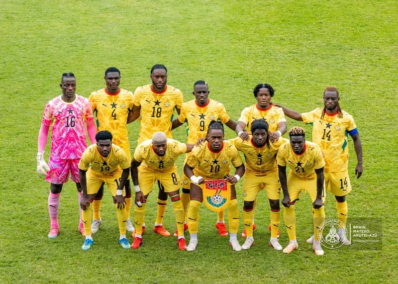 The Ghana national football team, the Black Stars, pose for a team photograph on a lush green pitch ahead of a match. The players are dressed in their yellow home kits with the iconic black star, while the goalkeeper stands on the left in a vibrant pink patterned kit.