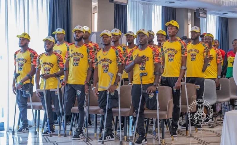 Members of Ghana's national amputee football team, the Black Challenge, stand together during an official event. The players are dressed in matching yellow jerseys and caps, many utilizing crutches as they appear to be observing a formal ceremony, likely the national anthem.