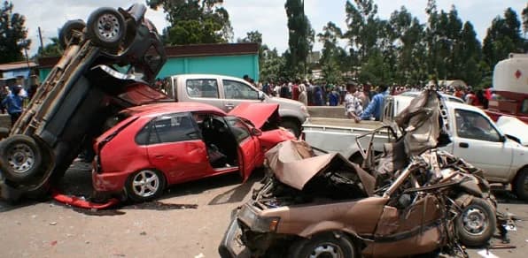 A multi-vehicle road traffic accident scene showing several severely mangled cars on a roadway. A dark vehicle is overturned and positioned vertically against a red hatchback, while a brown sedan in the foreground is completely crushed, with onlookers gathered in the background.