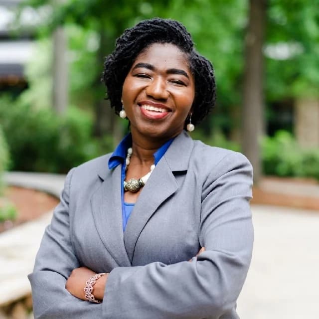 A professional portrait of Dr. Bertha Ayi, who is seen smiling with her arms crossed against a blurred outdoor background. She is wearing a grey blazer over a blue top, complemented by a beaded necklace and pearl earrings.