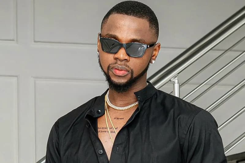A portrait of Nigerian musician Kizz Daniel wearing a black button-down shirt and dark sunglasses. He is adorned with several gold and diamond-encrusted necklaces and stands in front of a neutral-colored wall and metallic staircase railing.