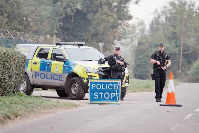 Two armed UK police officers stand guard next to a marked police vehicle and a 'POLICE STOP' sign on a rural road. The scene suggests a security checkpoint or an active investigation area, with one officer holding a rifle while a traffic cone cordons off the path.