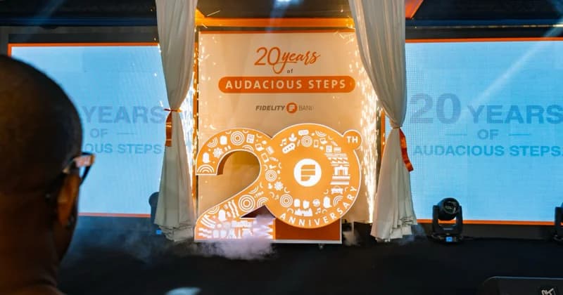 A festive stage setup for Fidelity Bank's 20th-anniversary celebration in Ghana, featuring a large orange '20' logo flanked by white curtains and digital displays. The scene includes indoor pyrotechnics with sparklers and a hazy atmosphere on the stage floor, creating a celebratory corporate environment.