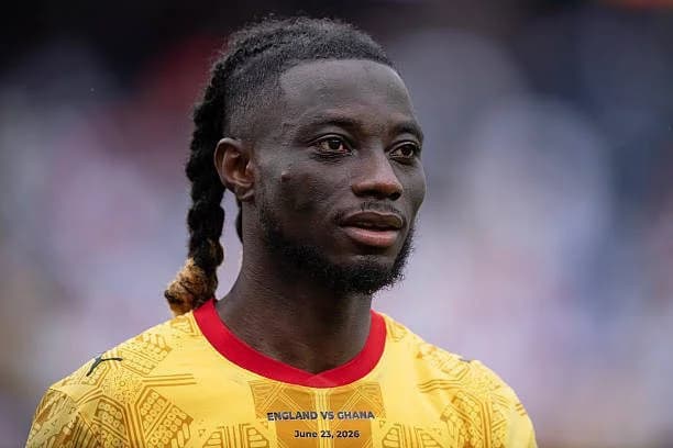 A headshot of Ghanaian footballer Gideon Mensah looking into the distance. He is wearing a yellow Ghana national football team jersey with red collar trim and decorative patterns, featuring text for a match against England.