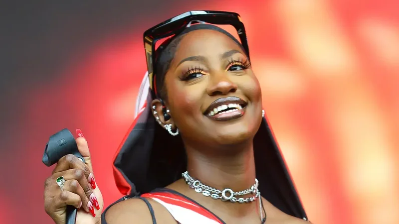 A close-up photograph of Nigerian singer Tems smiling and looking upwards during a stage performance. She is seen wearing stylish sunglasses on her head, silver necklaces, and elaborate nail art featuring a flame design while holding a microphone.