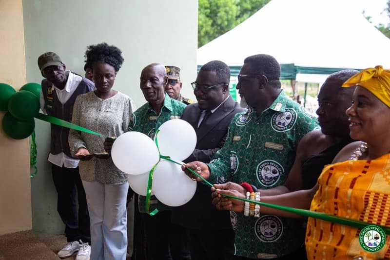 Officials and stakeholders are captured during a ribbon-cutting ceremony to inaugurate the Tree Crops Development Authority (TCDA) Bono Regional Office. Several individuals are wearing green shirts featuring the TCDA logo while holding a green ribbon decorated with white and green balloons.