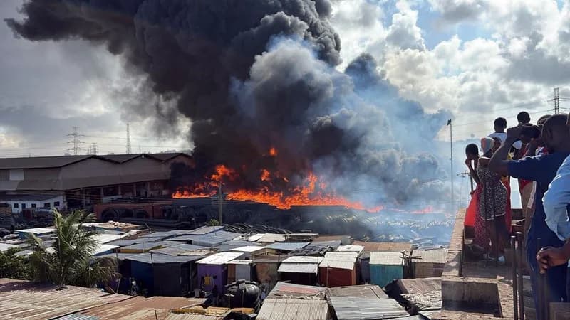 A massive fire rages at an industrial site, sending thick plumes of black smoke high into the sky. In the foreground, numerous corrugated metal structures are visible, while a group of onlookers watches the blaze from a nearby elevated platform.