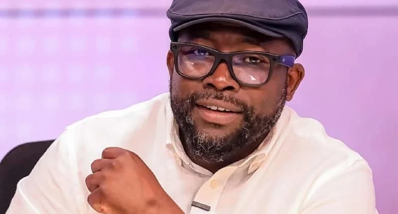 A close-up portrait of Senyo Hosi, a prominent Ghanaian economic and civic policy analyst. He is wearing a white shirt, dark-rimmed glasses, and a dark flat cap, appearing in what seems to be a television studio setting with a light purple background.