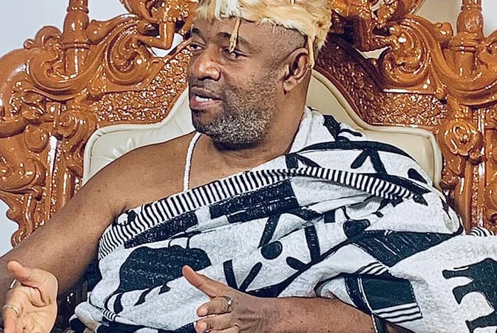 The Ga Mantse, King Tackie Teiko Tsuru II, is captured in a formal setting seated on an ornate wooden throne. He is dressed in traditional black and white patterned regalia with matching headgear, gesturing during a discussion or speech.