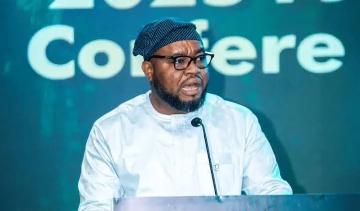 Isaac Sukparu, Member of Parliament for Sissala West, is captured speaking at a podium during a public event. He is wearing a white traditional garment and a patterned cap while addressing an audience behind a microphone against a teal backdrop.