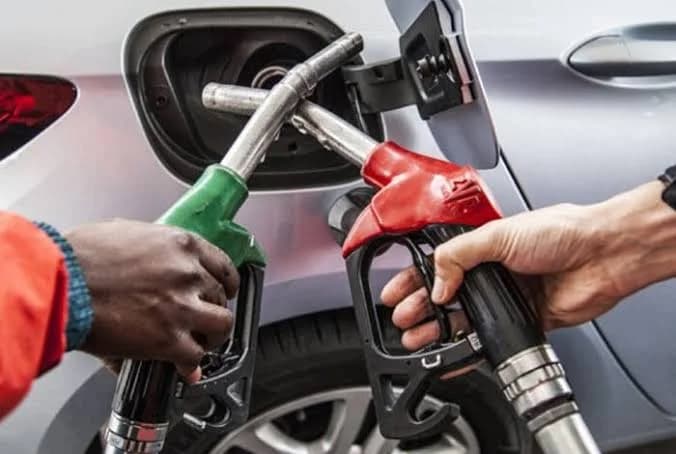 A close-up photograph showing two fuel pump nozzles, one green and one red, being held by two people near the open fuel tank of a silver car. The image captures the act of refueling at a petrol station, illustrating the retail side of the energy sector.