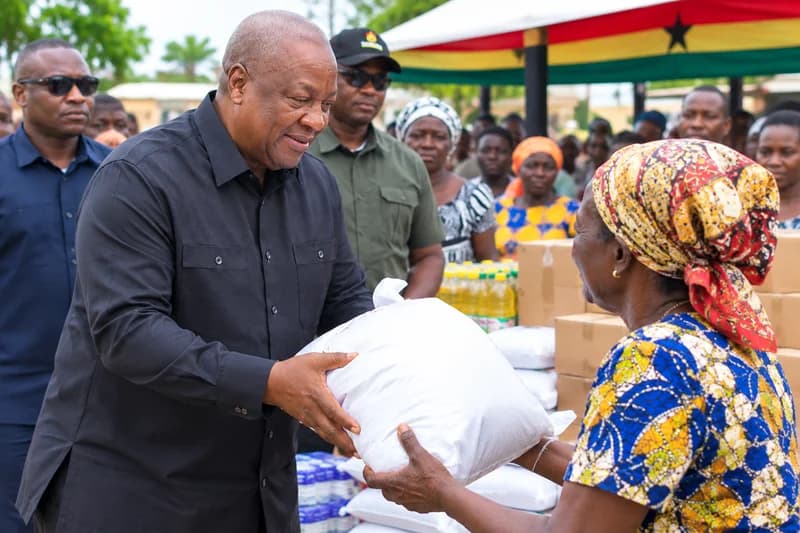 President John Dramani Mahama is pictured handing over a large white bag of relief supplies to an elderly woman. They are surrounded by a crowd and stacks of food boxes and bottled water under a canopy decorated with the Ghanaian flag.