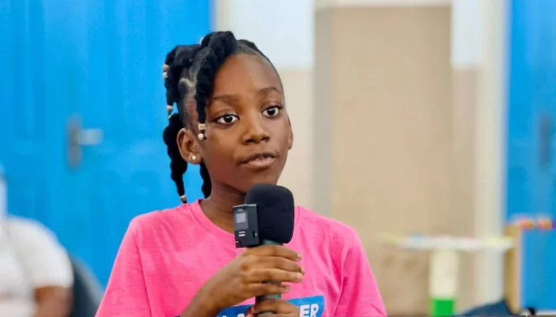 A young girl with braided hair and beads wearing a pink t-shirt speaks into a microphone during an event. The background features bright blue doors, and she appears focused as she addresses an audience, likely at the 2026 EARTH Summit.