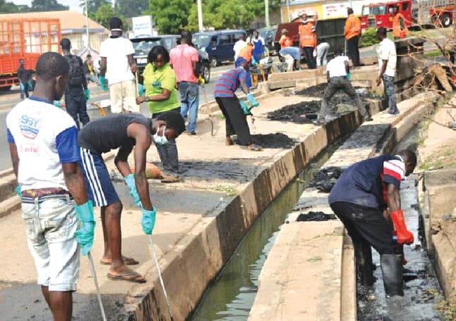 A group of people in Ghana are shown participating in a community cleanup exercise, specifically desilting a roadside concrete gutter. Participants are seen wearing protective gear like gloves and masks while using tools to remove debris, with a fire truck and other workers visible in the background.