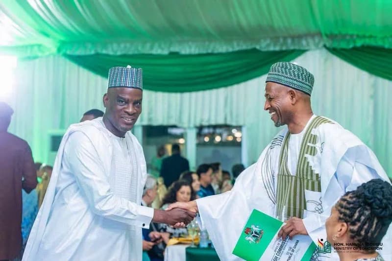 A photograph capturing a formal handshake between two men in traditional West African attire at a diplomatic event. The setting is decorated with green and white drapery, and one man holds a document bearing the Nigerian coat of arms, signifying a bilateral engagement between Nigeria and Ghana.