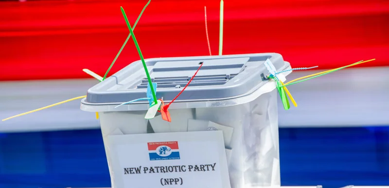 A translucent ballot box for the New Patriotic Party (NPP) is filled with ballots and secured with several colorful plastic seals. It is positioned against a background featuring the party's traditional red, white, and blue colors.