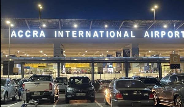A night-time photograph of the exterior of Kotoka International Airport in Accra, Ghana. The image displays several vehicles in a parking area with the main terminal building illuminated by bright white signage reading 'ACCRA INTERNATIONAL AIRPORT'.