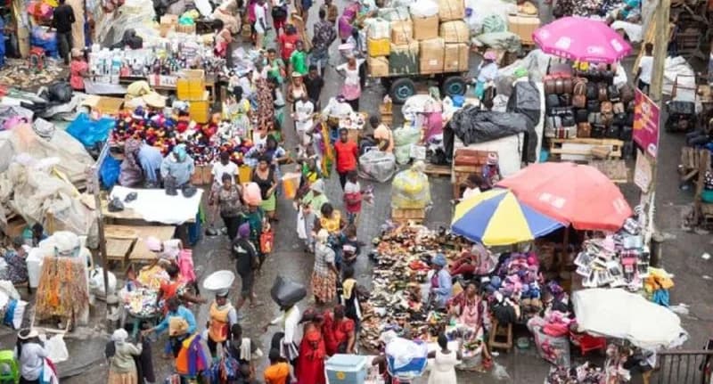 A high-angle view of a densely crowded market scene in Ghana, showing traders and shoppers occupying every available space on the ground. Colorful umbrellas, stacks of merchandise, and heavy pedestrian traffic highlight the severe congestion issues typically found in areas like Kasoa.