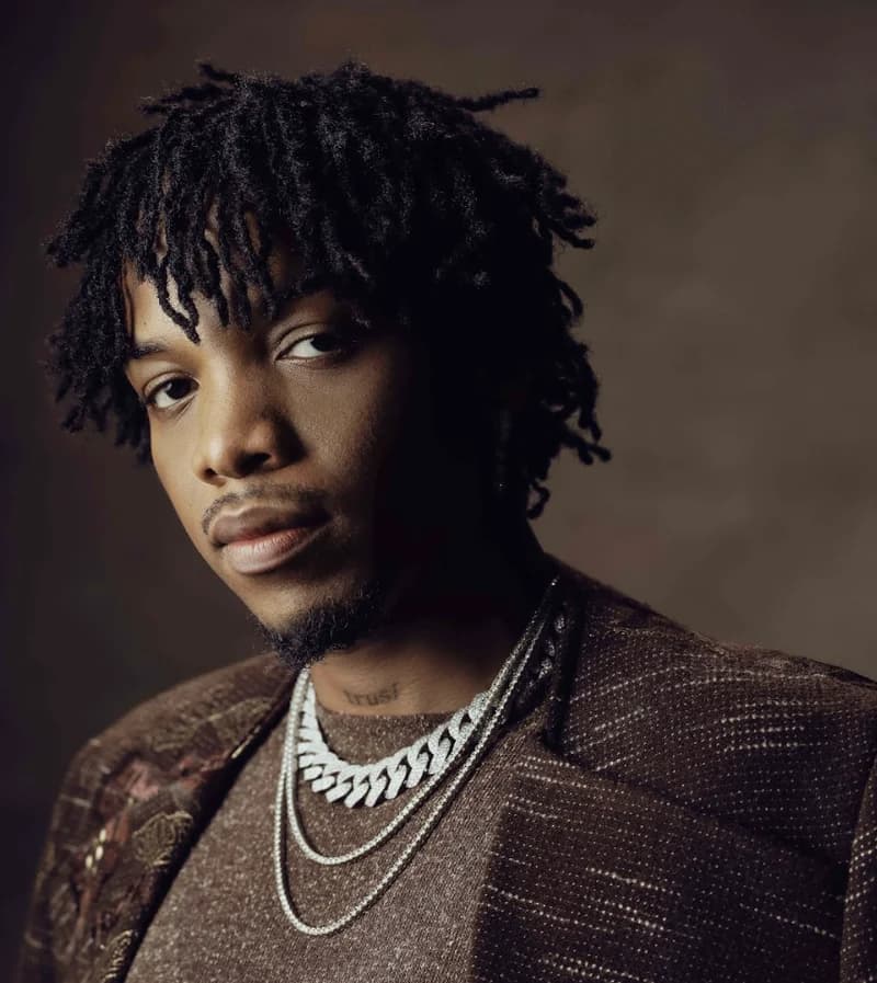 A professional studio portrait of the Nigerian musician Tekno (Augustine Miles Kelechi) against a dark, textured background. He is wearing a brown patterned jacket and silver jewelry, and a tattoo reading 'trust' is visible on his neck.