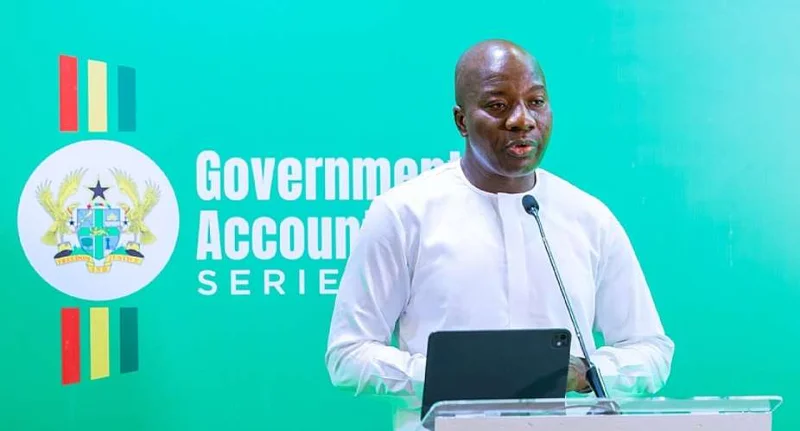 A photograph of a man in a white traditional shirt speaking at a podium during a 'Government Accountability Series' event. He is standing in front of a bright green backdrop that displays the Ghana Coat of Arms and national flag colors.
