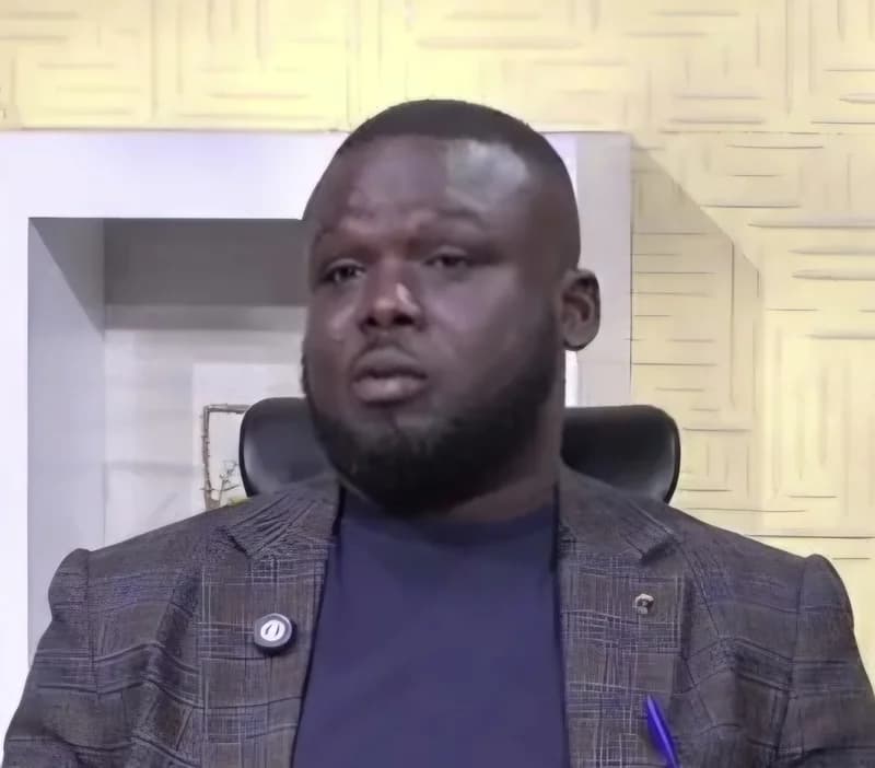 A man, likely a representative of the Ghana Maritime Authority, is seen in a studio setting during what appears to be a televised interview. He is wearing a grey patterned blazer over a navy blue shirt and is seated in a black office chair against a textured yellow-beige background.