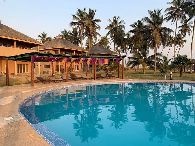 A serene view of a tropical hotel resort in Ghana, featuring a large circular swimming pool in the foreground. Several lounge chairs are arranged under a wooden pavilion next to villas and tall palm trees under a clear sky.