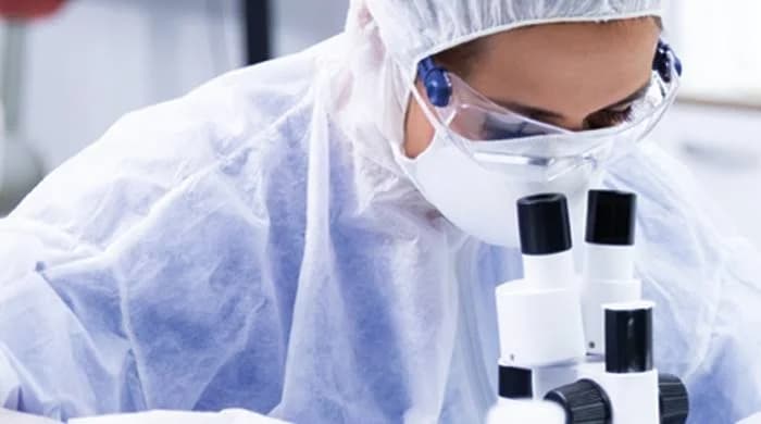 A laboratory researcher dressed in full personal protective equipment (PPE), including a white hazmat suit, face mask, and safety goggles, is shown operating a microscope. This close-up shot emphasizes the intense and precise nature of high-risk biological research in a clinical setting.