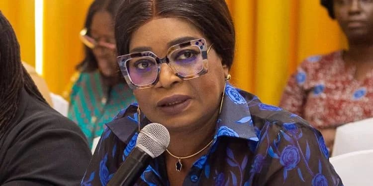 A close-up photograph of Honorable Lydia Alhassan, Member of Parliament for Ayawaso West Wuogon, speaking into a microphone during a formal meeting. She is wearing distinctive large-framed glasses and a blue floral shirt, with other attendees visible in the blurred background against yellow curtains.