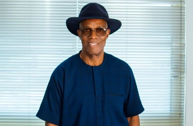 A portrait of Emmanuel Antwi-Darkwa, the Chief Executive of the Volta River Authority (VRA). He is wearing a navy blue short-sleeved shirt with a matching wide-brimmed fedora hat and tinted glasses, standing in front of horizontal window blinds.